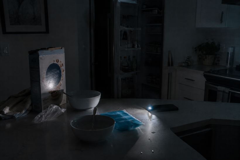 A dark kitchen lit by a phone flashlight, cereal box and bowls on the counter, fridge door open